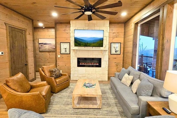 Rustic log cabin living room with stone fireplace, ceiling fan, leather and gray upholstered seating, and mountain views through glass doors.
