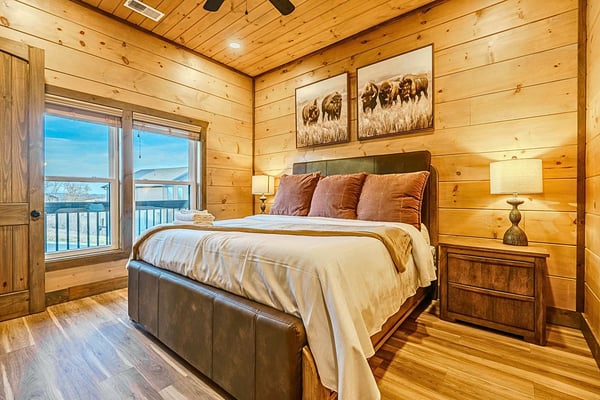 Open-plan cabin interior with wood ceiling, gray and tan seating, ceiling fan, kitchen area, and bedroom visible in background.